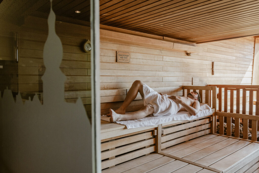 Ein Mann liegt mit einem Handtuch in einer Holzsauna und ruht sich auf der obersten Bank aus.