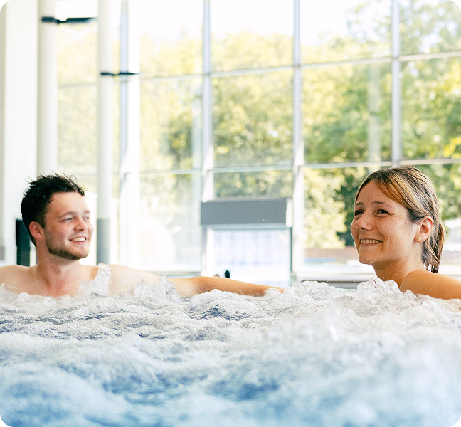 Mann und Frau sitzen lächelnd im sprudelnden Whirlpool eines lichtdurchfluteten Innenbereichs.