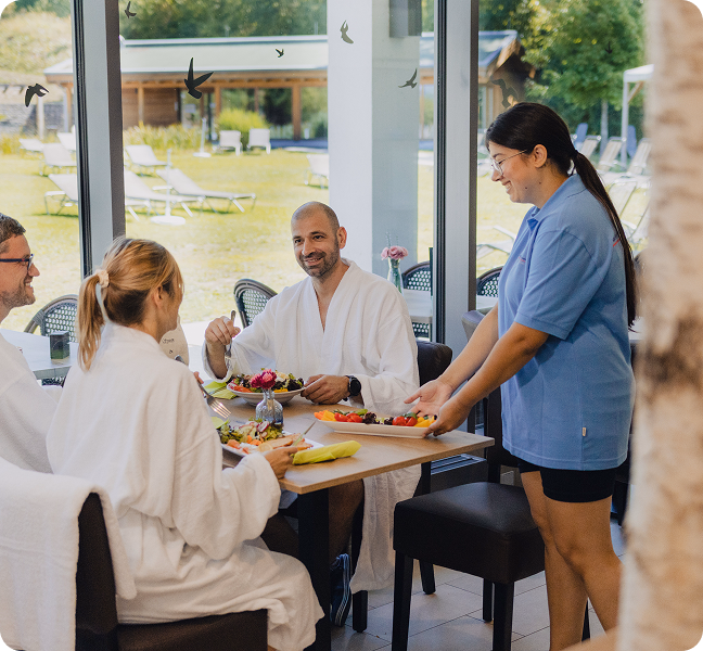 Gruppe von Menschen im Bademantel genießt frische Speisen im lichtdurchfluteten Bistro mit Blick ins Grüne.