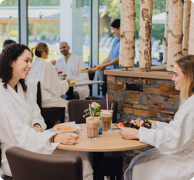 Zwei Frauen im Bademantel sitzen an einem Tisch im Bistro und genießen Speisen und Getränke, im Hintergrund weitere Gäste.