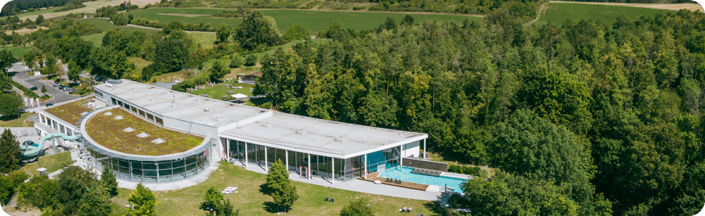 Luftaufnahme eines modernen Freizeit- und Erlebnisbads mit Flachdach, Außenschwimmbecken, großer Glasfassade und umliegender grüner Wald- und Wiesenlandschaft.