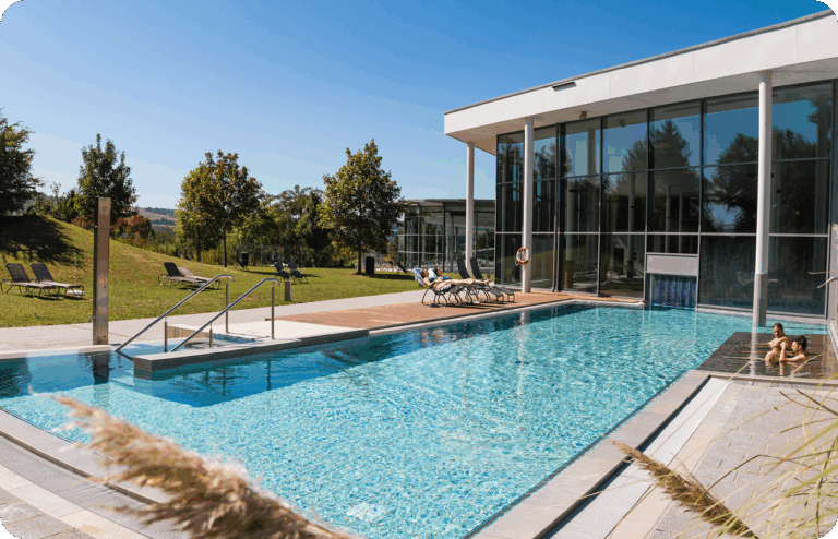 Außenpool mit Holzdeck und Liegen, umgeben von Bäumen und moderner Architektur bei strahlendem Sonnenschein.