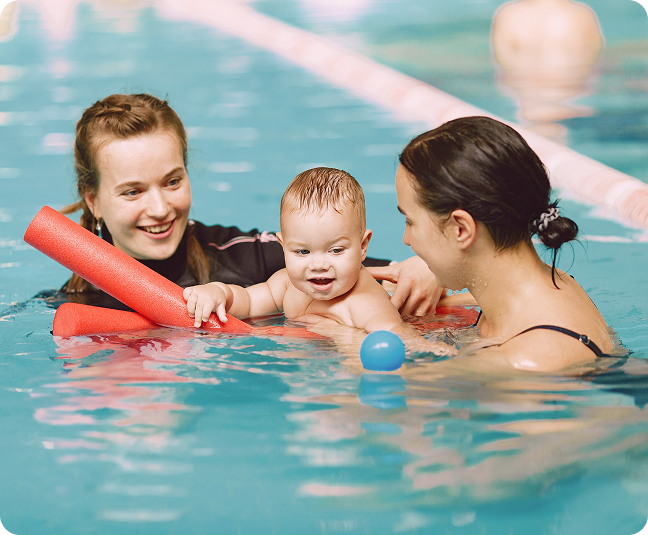 Ein Baby wird beim Schwimmkurs liebevoll von zwei Erwachsenen begleitet, spielt mit einer Poolnudel und einer kleinen blauen Kugel im Wasser.